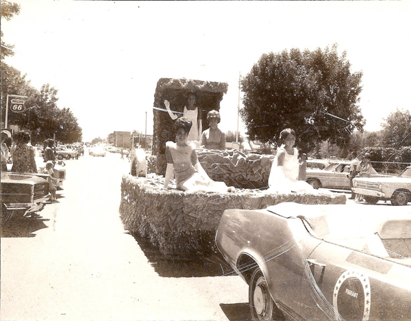 Parade float with three women in formal dresses sitting on it, moving down a street. A person stands and waves from a raised part at the back of the float. Several cars are parked along the street, and people are watching the parade from the sidewalks. A sign for "Phillips 66" is visible, along with another sign for "LELAND." A car with the word "PAGEANT" on its side is in the foreground. Trees are in the background.