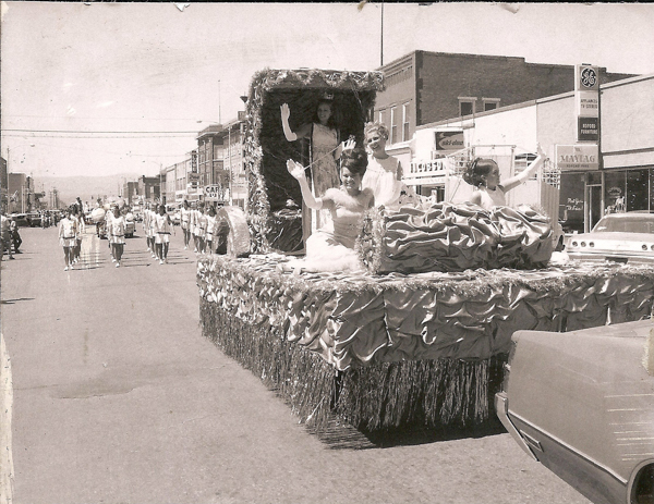 A parade scene on a street with several people riding on a decorated float. The float is adorned with patterned fabric and tinsel, with four women seated and waving. There is a group of people marching behind the float. Buildings line the street, with visible signs such as "CAFÉ," "GE," "Maytag," and "Kopford Furniture."