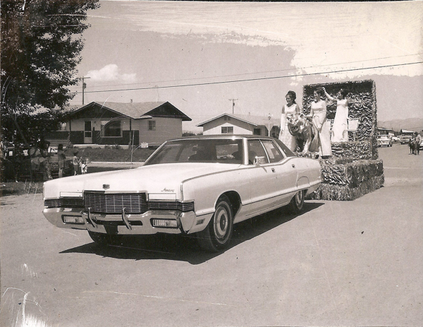 A car is driving in a parade, towing a decorated float. Three women in long dresses stand on the float, which features what appears to be a large model tiger. Houses and trees are visible in the background, and a small group of people is gathered along the roadside.