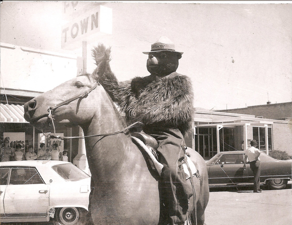 A person dressed in a bear costume rides a horse in a town setting. The costume has a hat with the word "SMOKEY" on it. In the background, there is a sign with the partially visible text "TOWN" and people standing near a storefront. Two cars are parked nearby, and a man is standing near one of them.
