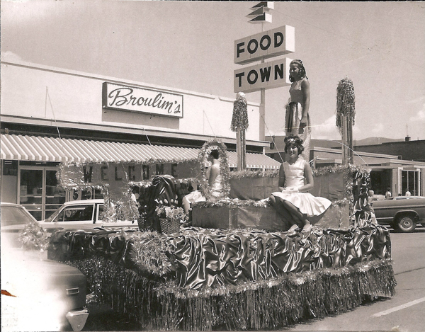 A parade float decorated with shiny fabric and tinsel features three women dressed in formal attire, one of whom is standing while the others are seated. A sign on the float reads "WELCOME." Behind them is a building with a sign reading "Broulim's" and another sign that says "FOOD TOWN." A car and part of a parking lot are visible.