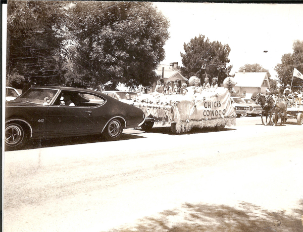 A parade with a classic car in the foreground, followed by a float carrying girls in dresses beneath a sign that reads "CHICK'S CONOCO." In the background, a horse-drawn carriage carries additional participants, with houses and trees lining the street.
