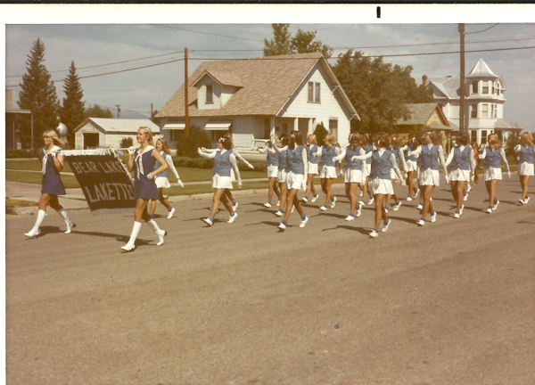 A group of young women in uniform march along a residential street. They are wearing blue vests and white skirts, with white gloves and boots. At the front, two women hold a sign that reads "BEAR LAKE LAKETTES." In the background, there are houses with lawns and trees, and the sky is clear.