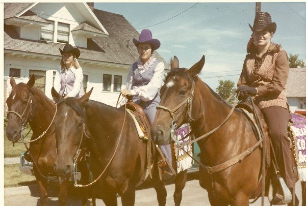 Three women are sitting on horses in front of a house, each wearing Western-style clothing and hats. They appear to be part of a parade; one horse is decorated with a cloth that has text on it, although the text is partly obscured. The house has white siding and a dark roof.