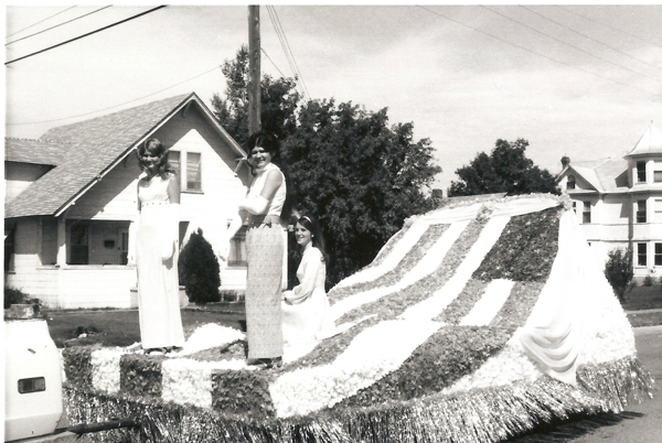Three women in formal dresses standing and sitting on a decorated parade float. The float is covered with floral patterns and stripes. Houses and trees are visible in the background, along with a pole and overhead wires.