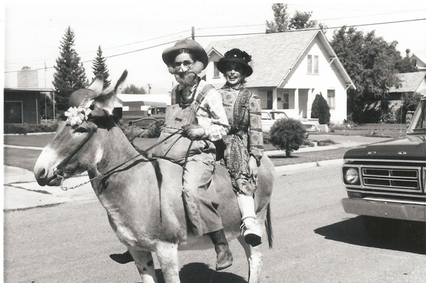 Two people riding a donkey on a residential street. The person in front is wearing a hat, glasses, and overalls, while the person behind is dressed in a patterned dress and hat. A car is partially visible on the right, and houses and trees line the street in the background.