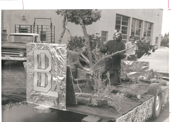 A person dressed in an animal costume stands on a parade float decorated with trees and foliage. Another person is visible in the background, smiling. The float has a large metallic decoration with a big letter "B" on it. A truck is parked nearby, and a building with large windows is in the background.