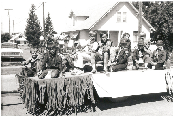 A group of boys in scouting uniforms sitting on a decorated float. They are in front of a house, and the float is adorned with hanging fringe and foliage. There are trees and houses in the background, as well as a parked car nearby.