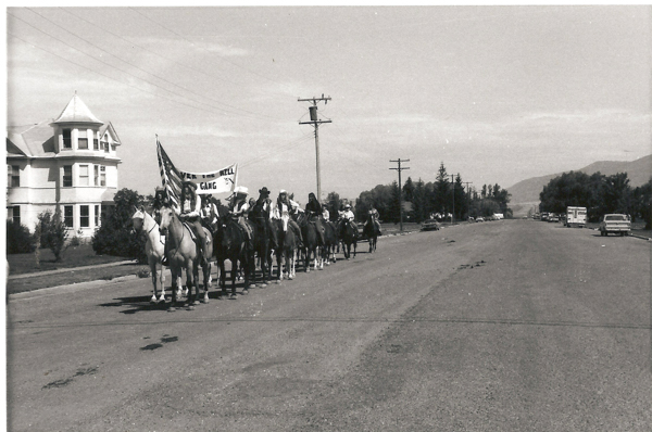 A group of people on horseback rides down a street. They carry a banner that reads "Over the Hill Gang." An American flag is also visible. The street has a large house on one side and a few cars parked along it. Trees and power lines are in the background.