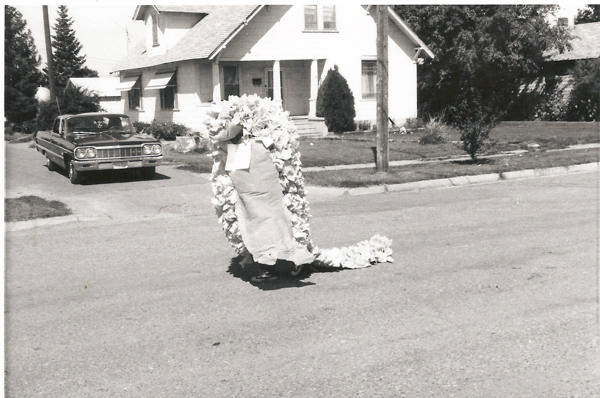 A person in a costume that looks like a large caterpillar or dragon, made of fabric and ruffles, is moving down a residential street. There is a vintage car parked on the side of the street, and a house with a porch and trees in the background.