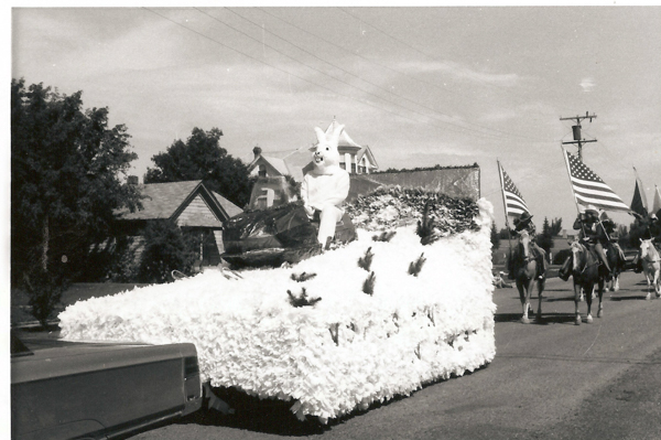 A person dressed in a rabbit costume sits on a float covered in decorative materials. The float is part of a parade with people on horses carrying American flags. Houses and trees are visible in the background.