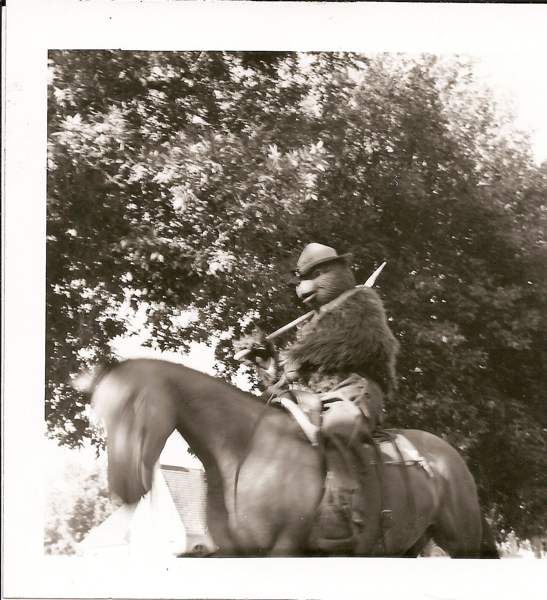 A person wearing a furry costume is sitting on a horse. They are holding a long stick or pole and wearing a hat. The background includes tree foliage and a building with a sloped roof.