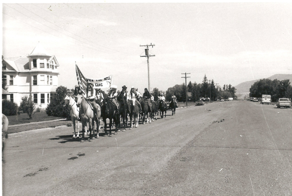 A group of people on horseback are moving down a wide road. They are carrying a banner that reads "OVER THE HILL GANG" and an American flag. Alongside the road is a large house and some parked cars. Utility poles and trees are visible in the background.