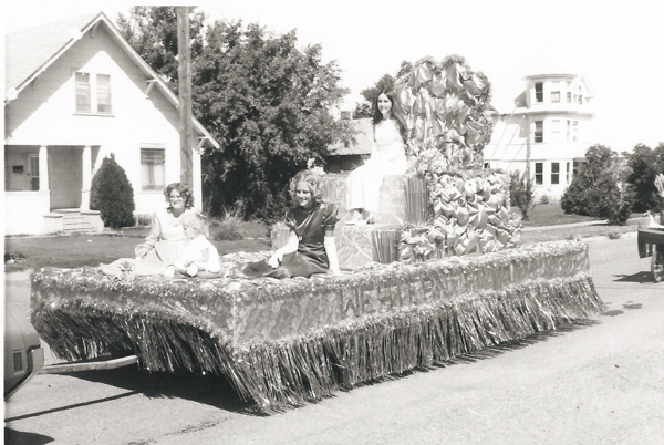 Three women sitting on a decorated parade float with fringes and ornamental designs. The word "WESTERN" is partially visible on the side of the float. They are in a residential area with houses and trees in the background.