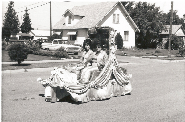 Two women sitting on a float decorated with draped fabric and flowers, riding on a residential street. Houses, a parked car, and trees are in the background.