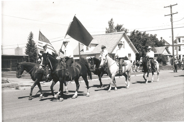Five people on horseback ride down a street, with a few holding flags. One carries an American flag, and another carries a plain flag. They appear to be participating in a parade. Several houses and a few spectators are visible in the background.
