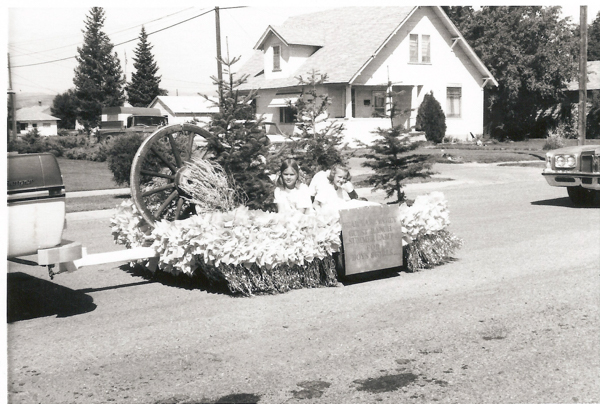 Two children are sitting on a decorated float being towed by a vehicle in a street. The float is adorned with flowers, small evergreen trees, and a large wagon wheel. A sign on the float reads "BUCKAROO BREAKFAST OUR SUNDAY STAMPEDE FLOAT BOYS AND GIRLS." Houses, cars, and utility poles are visible in the background.