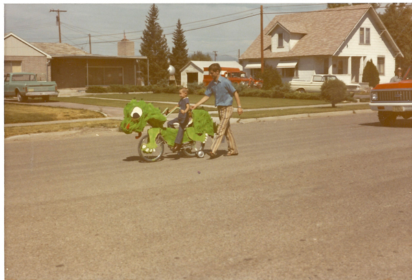 A child sitting on a bicycle decorated to look like a green dinosaur, complete with eyes and teeth. An adult is walking beside them, holding the rear of the bicycle. The scene takes place on a suburban street with houses, a green truck, and a red car visible in the background.