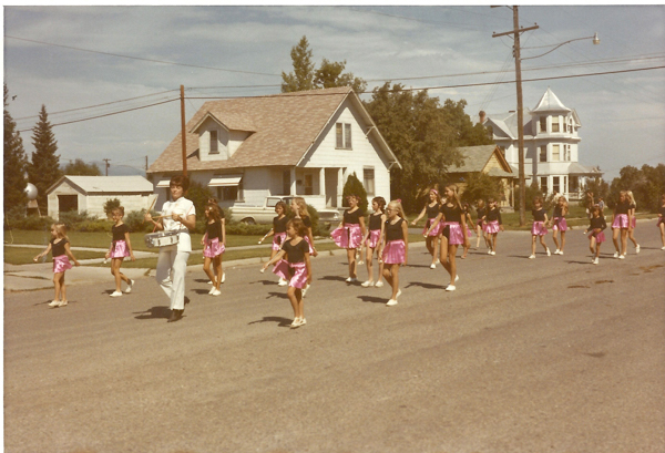 A group of young girls in matching black tops and pink skirts march down a residential street, followed by an adult playing a drum. There are houses and trees in the background, with a power line crossing overhead.
