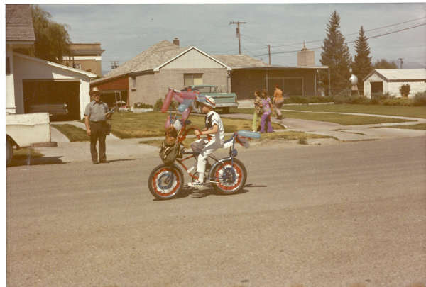 A child wearing a helmet rides a bicycle decorated with balloons and patriotic colors on a residential street. An older man stands nearby with a hat, holding an object. In the background, three people are walking on the sidewalk near some houses and trees.