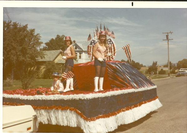 Three people are on a parade float decorated with red, white, and blue colors. They are wearing patriotic clothing with hats adorned with American flag patterns. Two are standing and holding small American flags, while one is seated on the float. A procession of American flags and decorations surrounds them. In the background, there are houses and trees lining the street.
