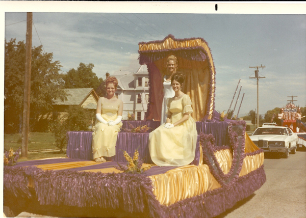 Three women in formal dresses and gloves are sitting on a decorated parade float. The float is adorned with purple and gold fabric, and it appears to be part of a parade. Behind them, a white car is visible, followed by another float with flags. Houses and utility poles are in the background.