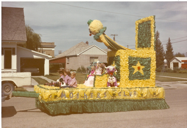 A parade float decorated with yellow and green flowers features a large clown head. Several children dressed in costumes sit on the float, with one child holding a stuffed animal. The float has the words "MOTHERS CLUB" displayed on its side. Residential houses are visible in the background.
