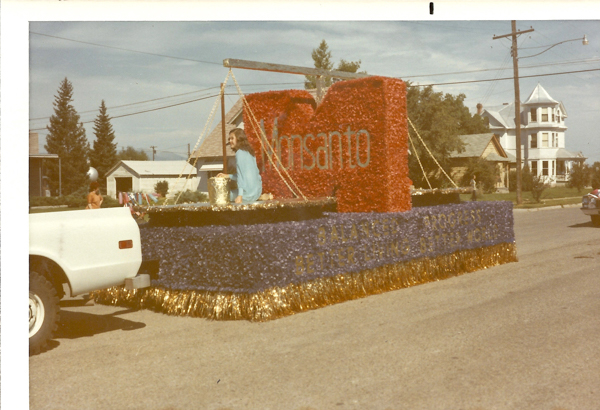 A person is sitting on a parade float decorated with red and blue floral patterns. The float has a large red letter "M" with the word "Monsanto" on it. The text "BALANCED PROGRESS BETTER THINGS FOR A BETTER WORLD" is visible on the side. The float is attached to a white vehicle. Houses and trees line the street in the background.