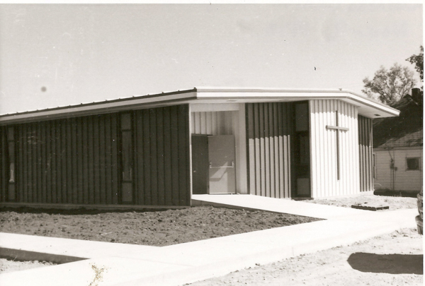 A single-story building with a modern design featuring vertical paneling. A large cross is mounted on one of the exterior walls. A pathway leads to the entrance of the building. A small house is visible in the background on the right side.