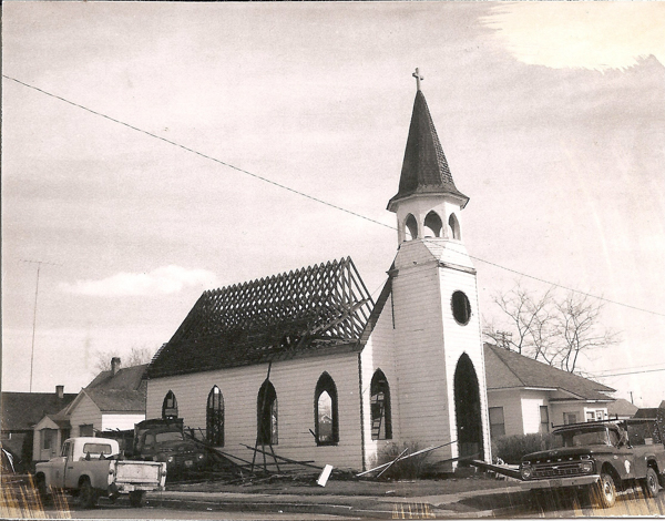 A church building with roof under construction, featuring a tall steeple with a cross on top. Several trucks are parked around the church. Nearby, there are small houses and leafless trees. Broken wooden structures and debris are visible around the scene.