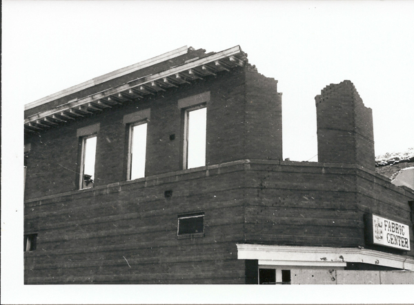 A partially demolished brick building with three tall windows. The top part of the structure shows damaged corners. A sign on the lower right side reads "Fabric Centre" with a partially visible logo.
