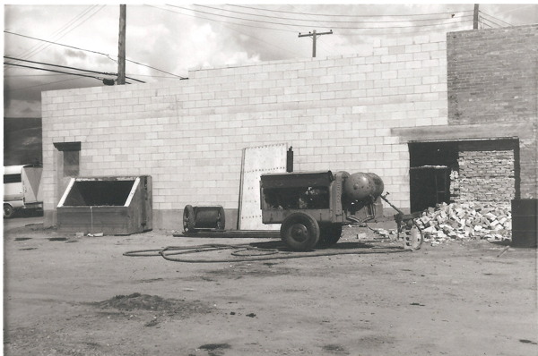 A construction or industrial site showing a large machine with wheels and a spherical component, hoses lying on the ground, and a pile of bricks or debris. A cinderblock building and a vehicle are present in the background, along with power lines.