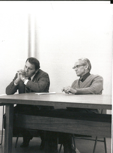 Two men seated at a table. One is resting his chin on his hand, appearing thoughtful, while the other looks ahead with arms resting on the table. The setting appears to be a simple room without decoration.