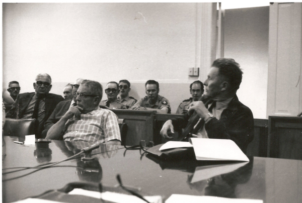 A group of men seated in a room. In the foreground, two men sit at a table, one in a checkered shirt and the other holding a pen or microphone. In the background, several men in uniform and glasses are seated in a row, looking towards the front of the room. There are papers on the table, and a microphone is visible.