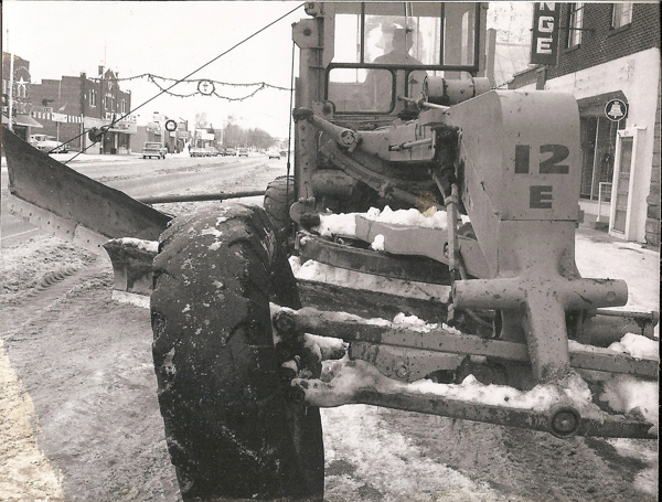A large vehicle equipped with a snowplow is parked on a snow-covered street. The vehicle's side shows the text "12 E." In the background, there are several buildings and parked cars. Decorations are hung across the street above the roadway.