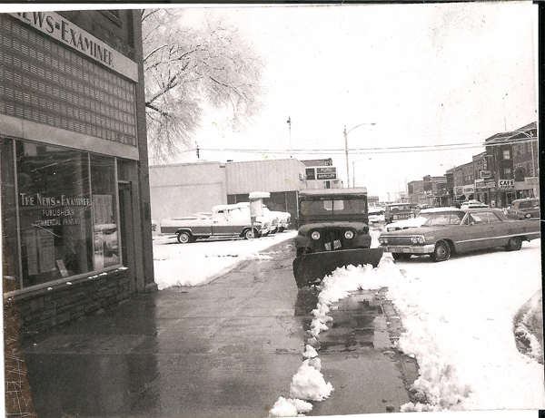 A snowy street scene featuring cars parked along the road. On the left, a building displays a sign reading "THE NEWS-EXAMINER" with additional text "PUBLISHERS" and "COMMERCIAL PRINTING." A pickup truck and a car are visible, partially covered in snow. In the background, a sign for "FORD MERCURY" is seen, and numerous other buildings and storefronts line the street, including one labeled "CAFE." Snow covers the sidewalk and parts of the road.