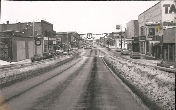 A snow-covered street with cars parked on both sides. Buildings line the street with various signs, including "Standard," "Chevrolet," "Buick," "Loans," and "Bar." Holiday decorations hang across the street. The road has visible tire marks, and snow piles up along the edges.