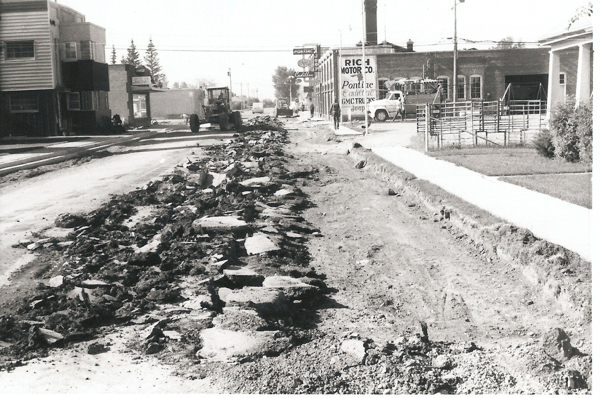 A street undergoing construction with rough gravel and debris scattered across it. On the left, there are buildings, one with a "Rich Motor Co." sign. A tractor is visible on the road, and a few cars are parked. A sidewalk and grassy area are on the right. Several signs for businesses are in the background.