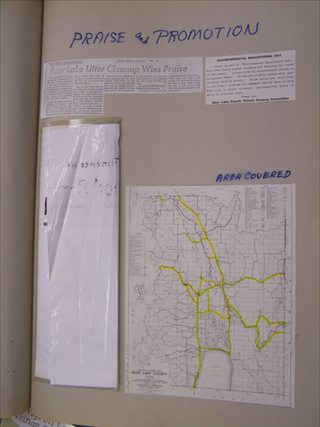 A scrapbook page titled "Praise and Promotion" features a newspaper clipping with the headline "Bear Lake Litter Cleanup Wins Praise," describing the involvement of students in cleaning efforts. There is a notice about "Environmental Educational Day" by the Bear Lake County School Cleanup Committee, encouraging responsible garbage disposal and community pride. Adjacent to these is a map titled "Bear Lake County," with certain routes highlighted, under the heading "Area Covered." A printed article is encased in a plastic sleeve.