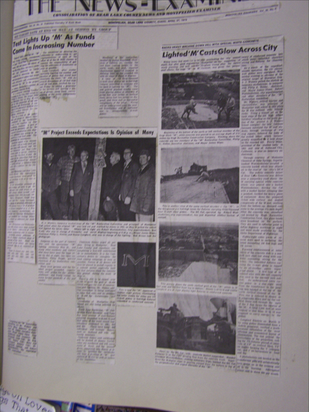 A newspaper page with multiple columns of text and several photographs. The header reads "The News Examiner" and features articles titled "Test Lights Up 'M' As Funds Come In Increasing Number" and "'M' Project Exceeds Expectations Is Opinion of Many." Another titled "Lighted 'M' Casts Glow Across City" includes three images: a group of people standing with a post, individuals working on a large letter "M" project, and the letter "M" lit up. The page also has smaller cut-out segments pasted onto it.