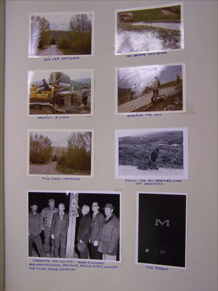A collection of seven images with handwritten captions. 

The first image shows a dirt road lined with trees leading to a hillside captioned "One leg complete." 

The second image shows a mix being spread on the ground, captioned "The Before and After."

The third image depicts a man operating machinery next to a mixer, captioned "Dropping a load."

The fourth image shows individuals spreading a mix on a sloped path, captioned "Spreading the mix."

The fifth image features a scene similar to the first image but further along, captioned "Two legs complete."

The sixth image shows a person kneeling and working on the ground, captioned "Fixing the all weather lite, Les Lanfrod."

The seventh image features a group of men standing near a wooden post at night, captioned "Throwing the switch - Alma J. Winters; Bob Woolstenhulme, Don Hunt, Mayor Olsen, Winters, Phil Hulme, Duane Langford."

The last image is a dark background with an "M" prominently displayed, captioned "The Result."