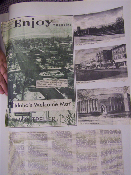 A hand holds a page from a magazine titled "Enjoy magazine." The main image shows an aerial view of a town with text that reads: "'Idaho's Welcome Mat' Montpelier." To the right, there are smaller images labeled "Bear Lake Memorial Hospital," "Wide Washington Street," and "LDS Stake Tabernacle." Below are blocks of text in columns.