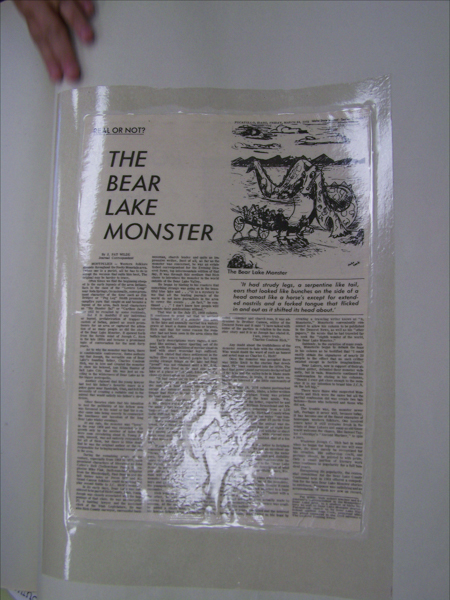 A hand is holding a laminated newspaper article labeled "REAL OR NOT? THE BEAR LAKE MONSTER" by J. Pat Wilde. The article features an illustration of a large, serpentine creature with a long tail and various animal-like features emerging from a lake, alarming a group of people in a horse-drawn carriage. The text describes the legend and sightings of the Bear Lake Monster.