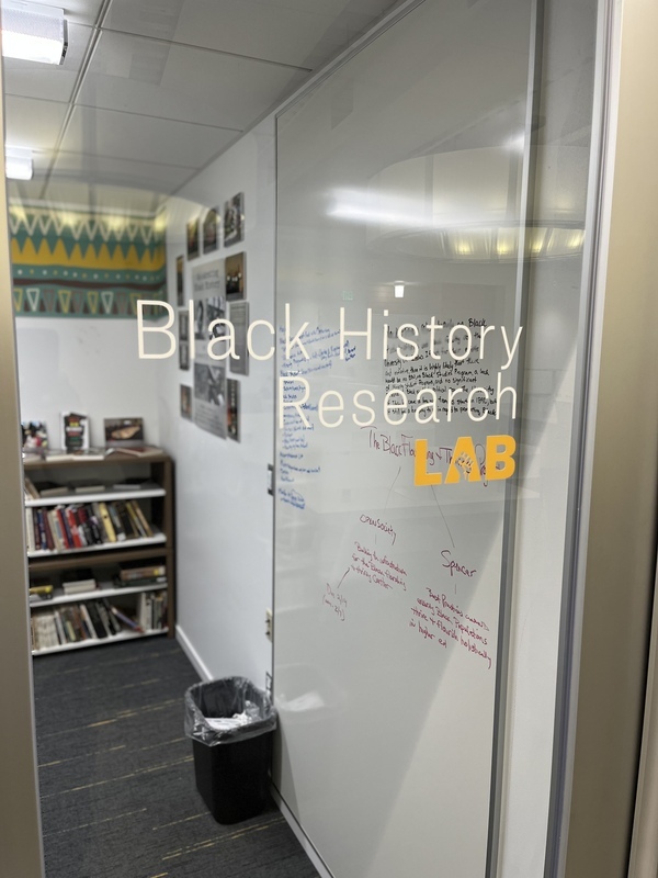 A view of the exterior window of the Black History Research Lab. Vinyl lettering reads "Black History Research Lab." Through the window, a whiteboard, bokoshelf, and posters can be seen in the workspace. 