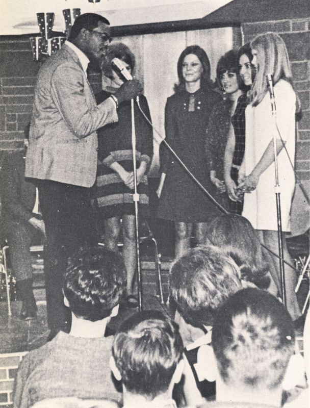 Joe Tasby standing on a stage and near a microphone, acting as an emcee for the Holly Frolics,  caroling group during Holly Week. 