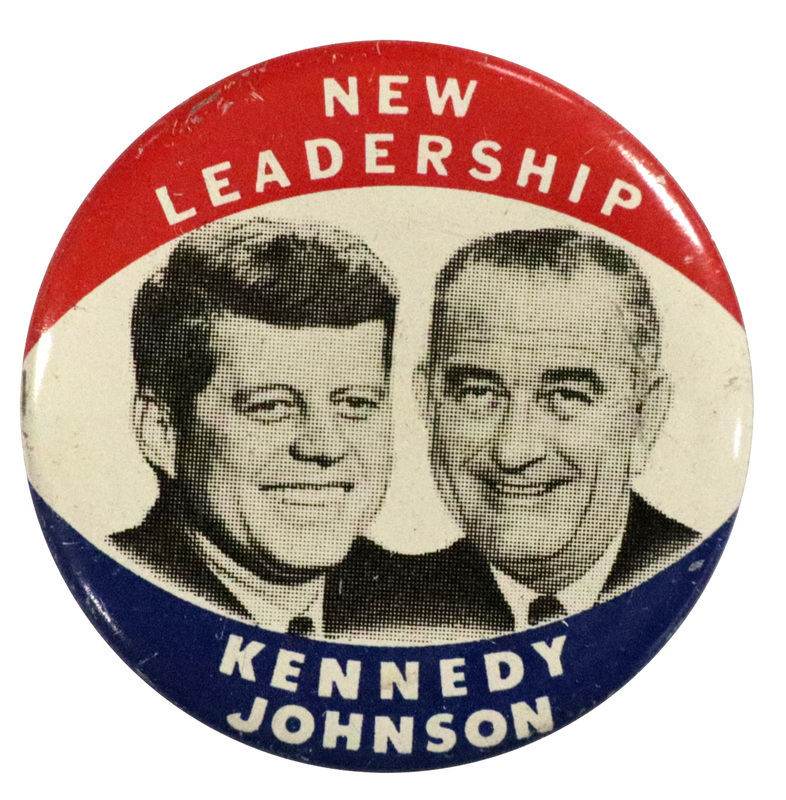 Round, white button with an accented red and blue border across the top and bottom. There are two grayscale portraits of John F. Kennedy and Lyndon B. Johnson at the center. White text reads "New leadership Kennedy Johnson". 