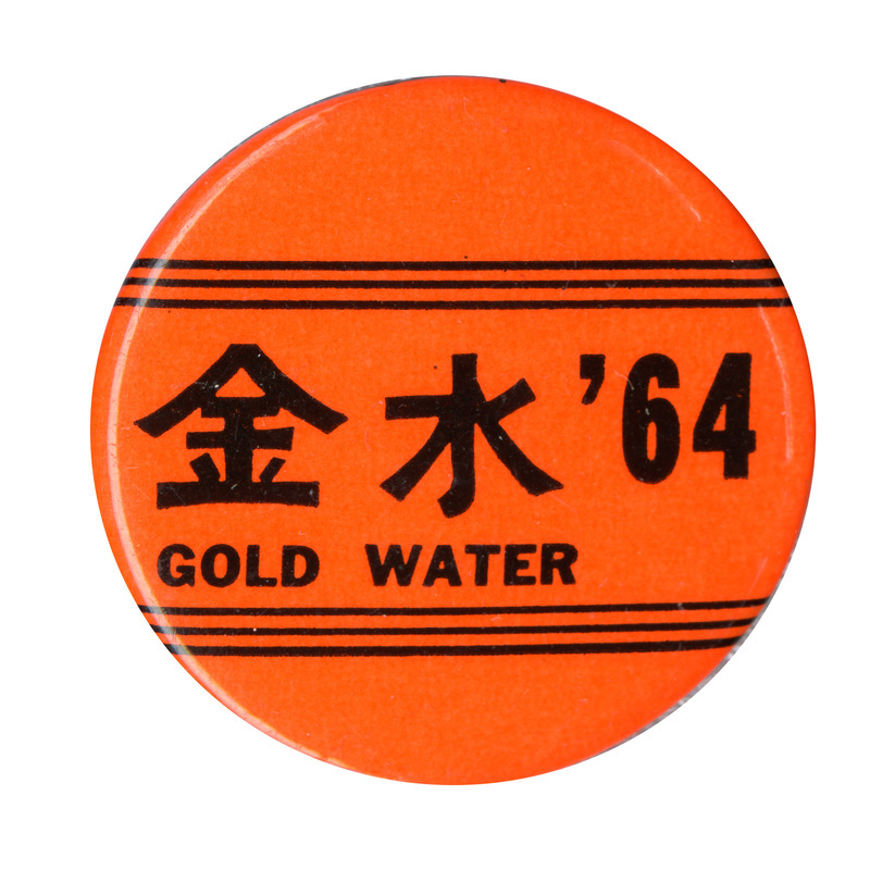 Round, orange button with narrow horizontal black lines. The text reads "Gold Water '64" and is accompanied by the Chinese characters for "gold" and "water".