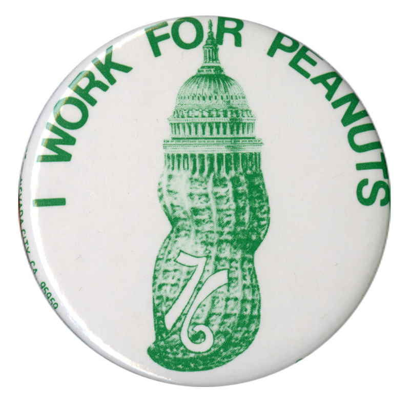 Round, white button with a green image of a peanut topped by the dome of the US Capitol. Green text reads "I WORK FOR PEANUTS 76"
