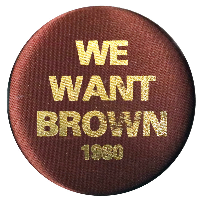 Round button covered in brown fabric. Shiny gold text reads "WE WANT BROWN 1980"
