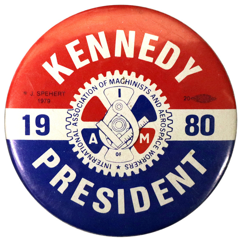 Round, red and blue button with a white band across the center. In the middle is a red, white, and blue drawing of measuring instruments against a gear that includes the text "INTERNATIONAL ASSOCIATION OF MACHINISTS AND AEROSPACE WORKERS I A OF M." White and blue text around the gear reads "KENNEDY 1980 PRESIDENT"